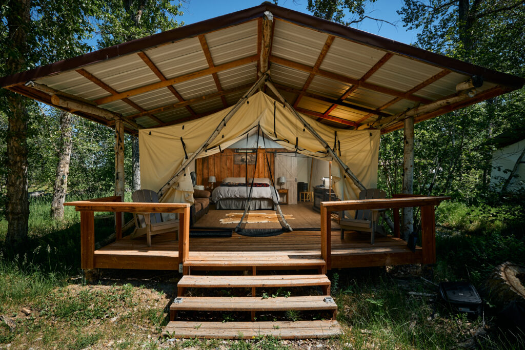 Exterior of a canvas-wall glamping tent under a metal-roofed pavilion, with a covered deck, steps, and porch chairs.