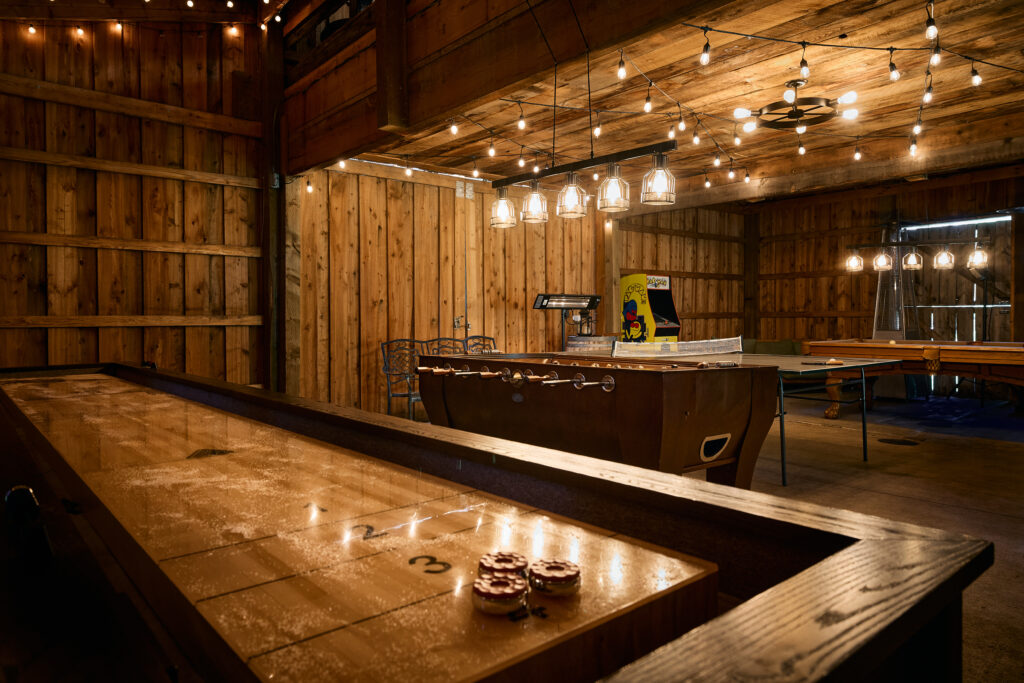 A wooden shuffleboard table in the game barn, with a foosball table and arcade cabinet lit by string lights