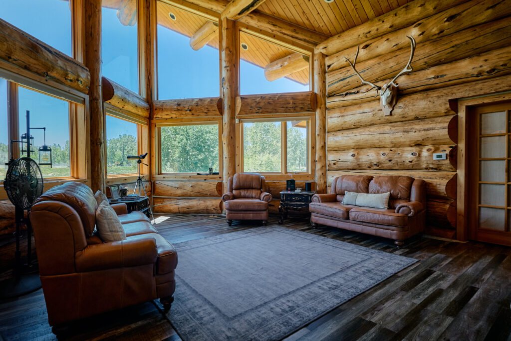 Great room with brown leather sofas and armchairs beneath soaring log-cabin windows and mounted antlers