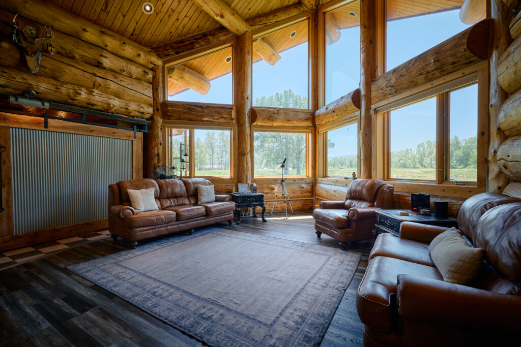 Leather sofas and armchairs around a rug in the great room, with tall windows framing open meadow and trees outside.