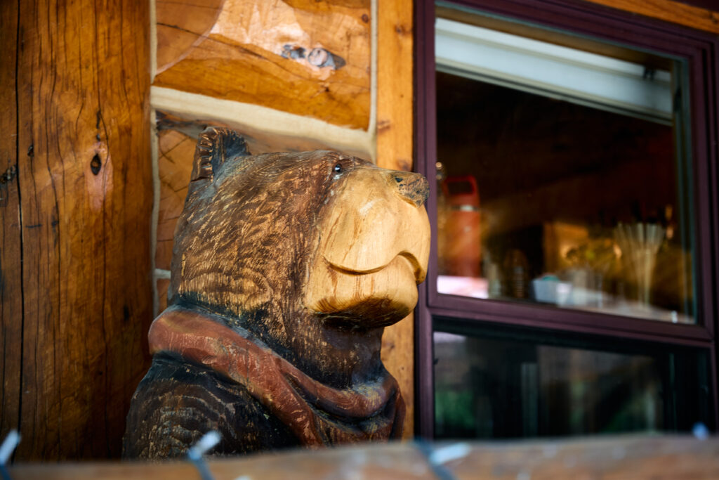 Carved wooden bear bust wearing a rust-red scarf gazes toward a maroon-framed lodge window