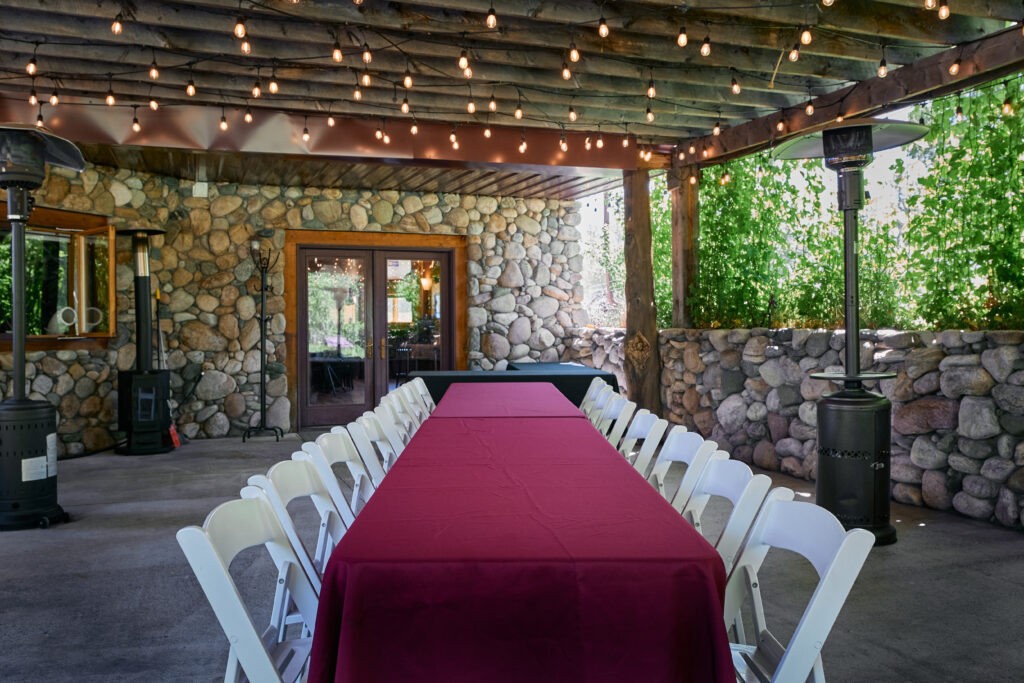 A covered patio strung with lights and hop vines, set with a long table in burgundy linens and white chairs.