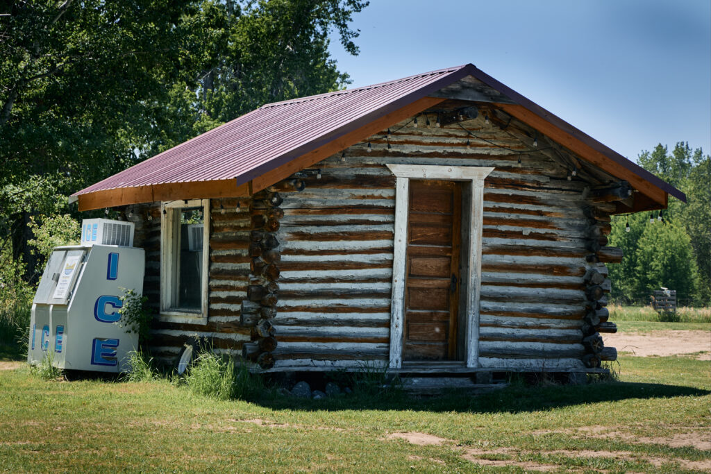 A weathered log cabin with a dark red metal roof and a vintage ICE vending machine outside on the property grounds.