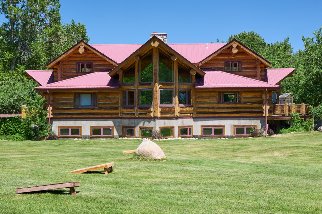 Front view of the lodge's log gables and great-room windows, with cornhole boards and a boulder on the lawn