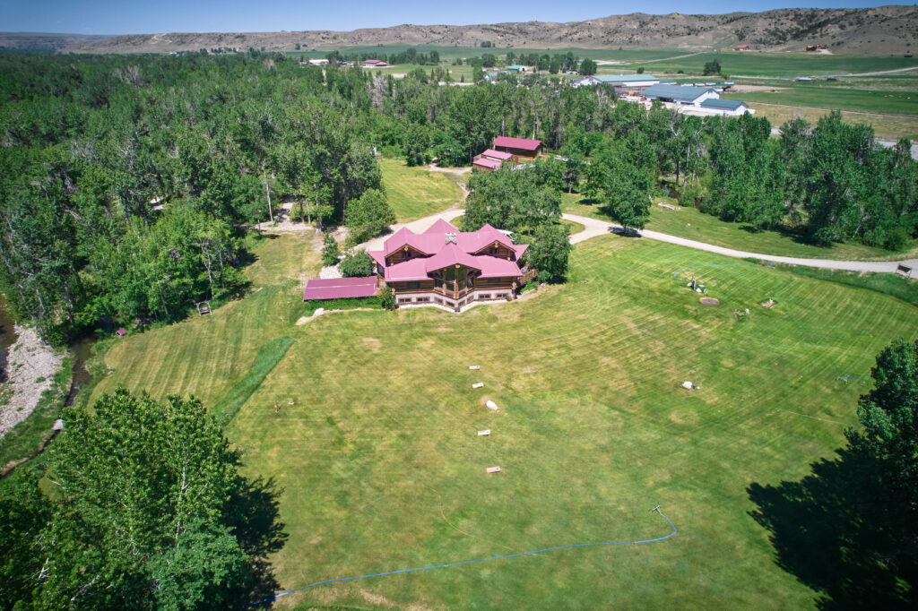 The maroon-roofed lodge and matching outbuilding sit amid cottonwoods far below, with a creek bend and foothills beyond