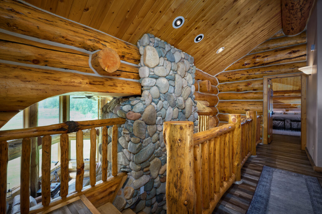 An upstairs hallway landing with a river-stone chimney and log railings overlooks the great room below