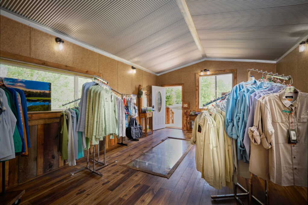 Rows of clothing racks fill the lodge's fly shop, with a glass floor panel and open door to the deck