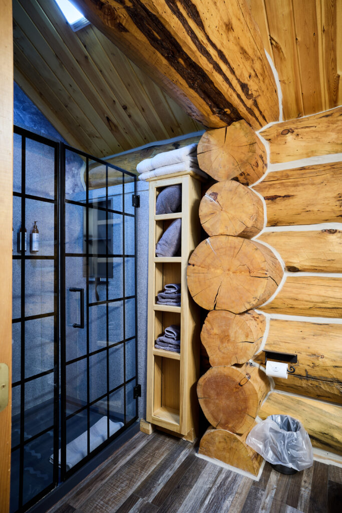 A glass-enclosed shower in a log-walled bathroom, with folded towels stacked on a built-in shelf