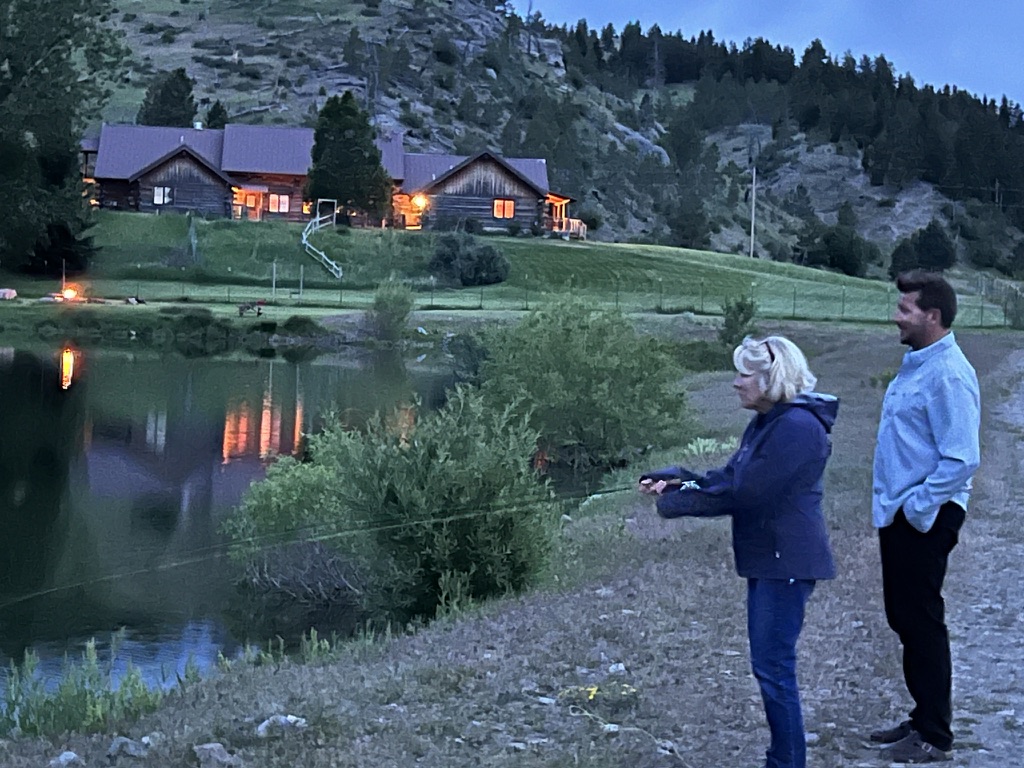 Two anglers fly casting on the trout pond at dusk with the lit lodge reflected in the water